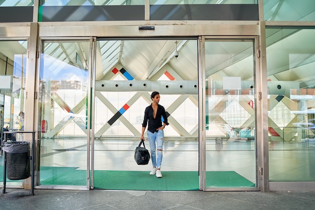 how to pick a sliding door, Indian man in black shirt and ripped jeans entering a modern building with a bag, geometric design in the background.